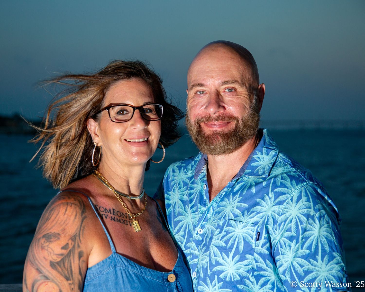 A smiling couple poses outdoors at dusk, woman with tattoos and glasses, man in tropical blue shirt.