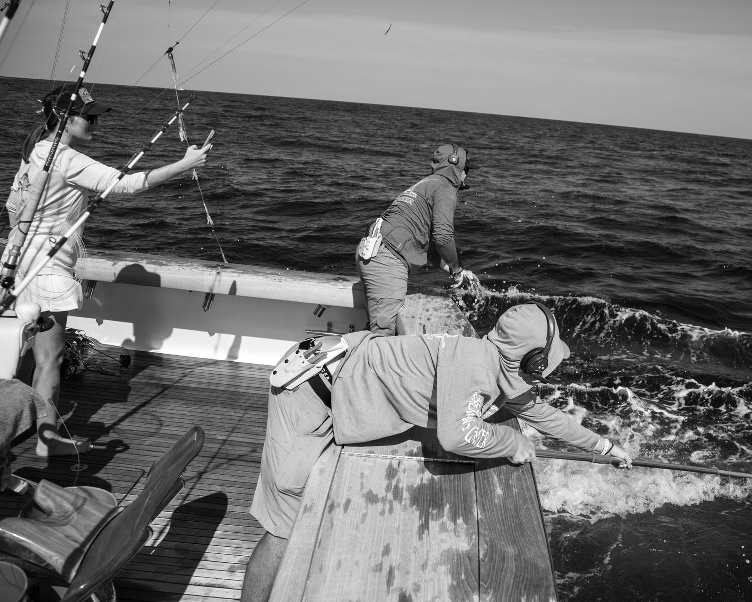 Three men fishing on a boat in open ocean, captured in black and white photography.