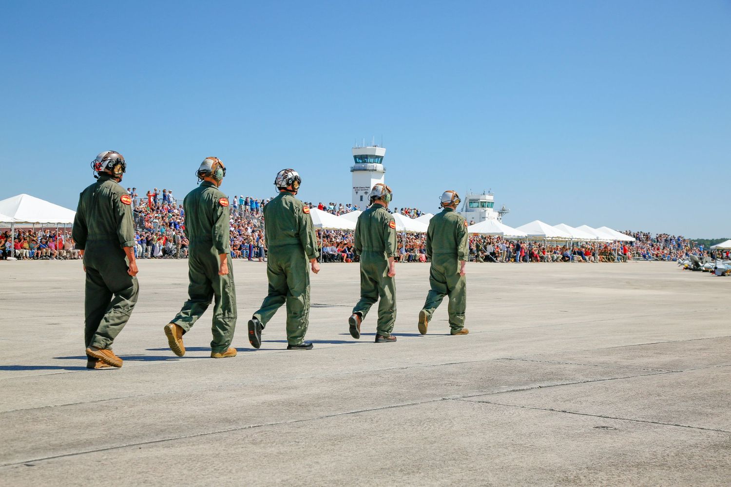 Military pilots in green flight suits march across an airfield tarmac at a crowded air show event.