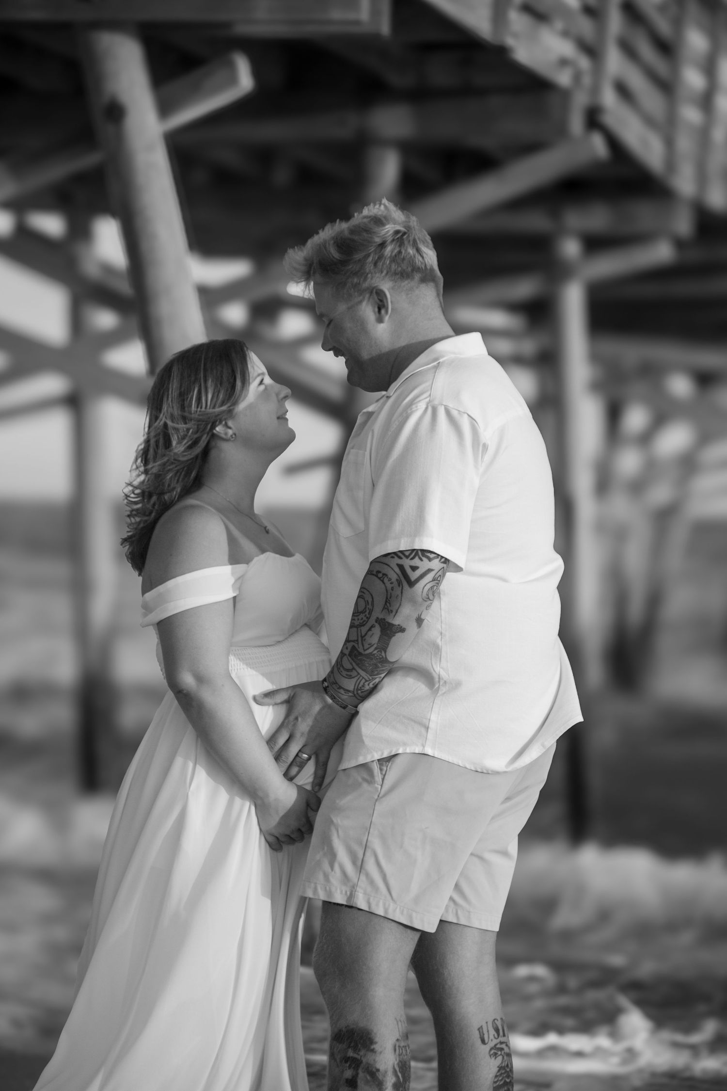 Black and white photo of a tattooed couple holding hands and gazing at each other under a beach pier.