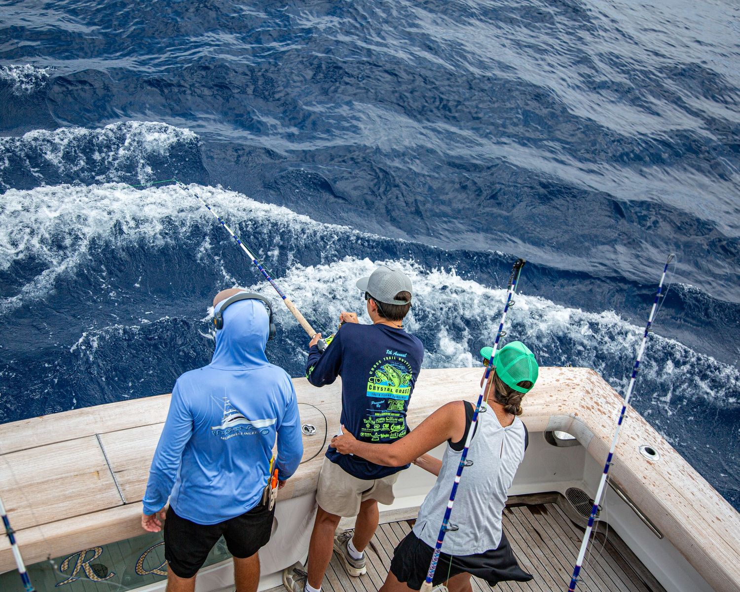 Three anglers fishing off the stern of a sport fishing boat on deep blue ocean waters.