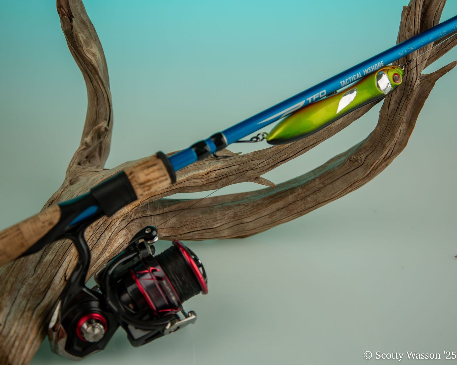 Blue fishing rod with spinning reel resting on driftwood against teal background.