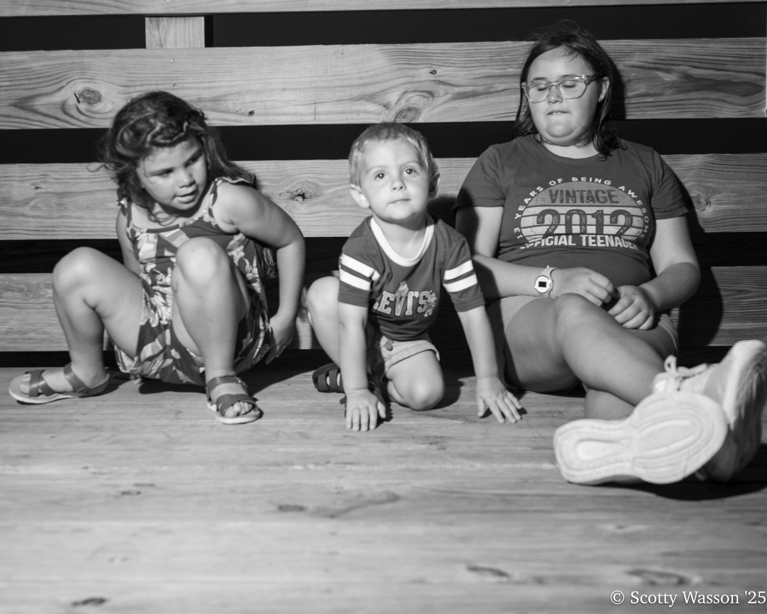 Black and white photo of three children sitting on wooden floor, two girls and a boy posing casually indoors.
