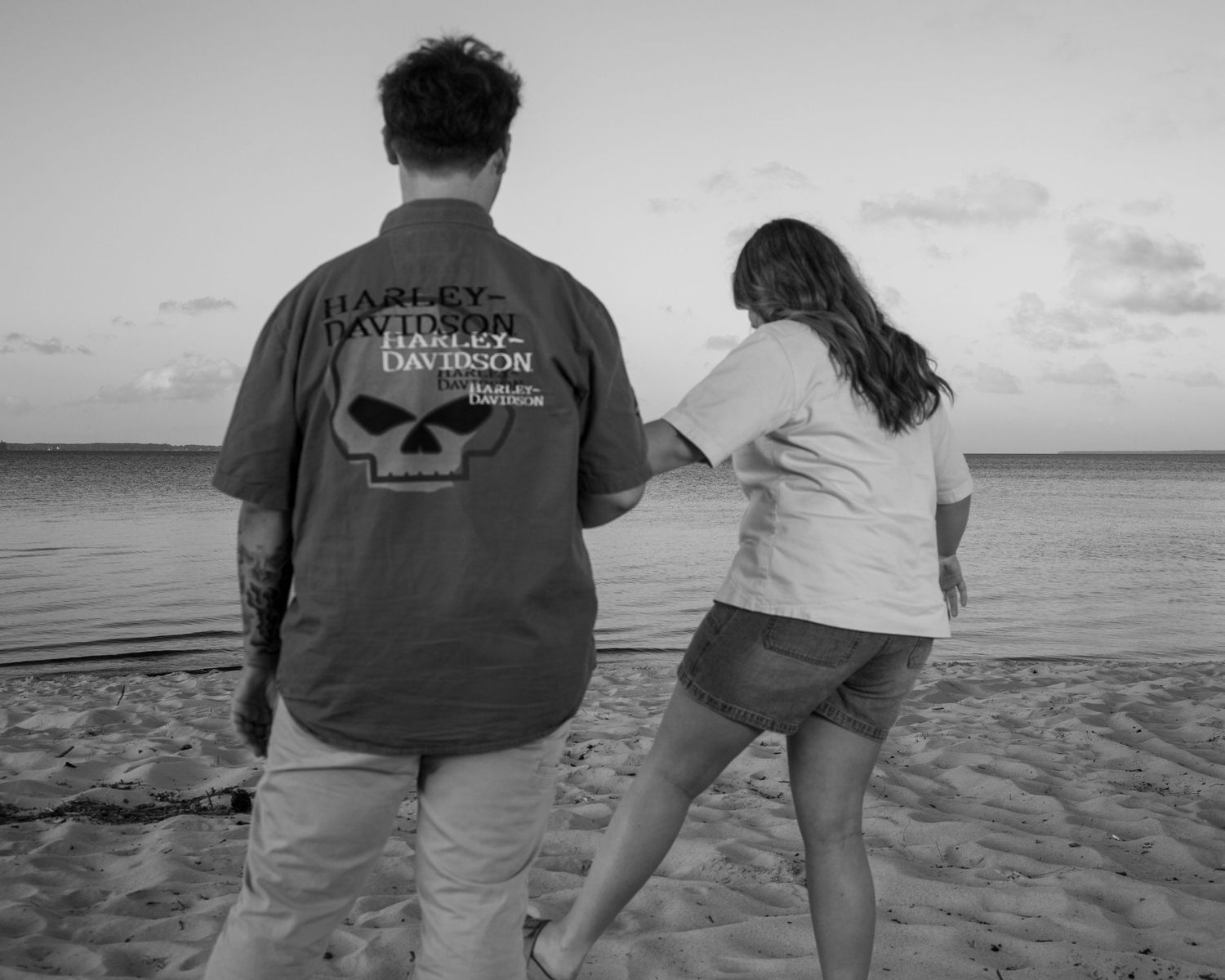 Couple walking on beach, man wearing Harley-Davidson skull tee, black and white photo.