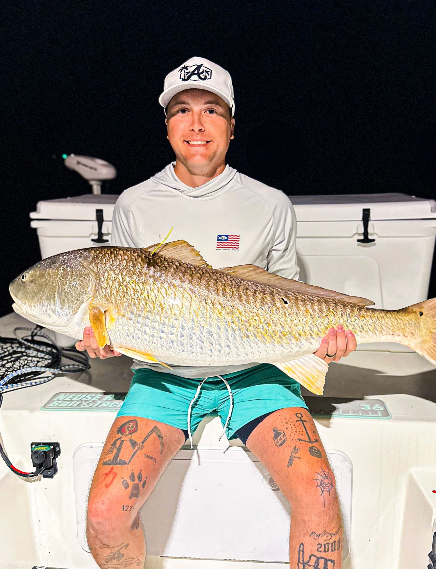 Man in white cap and teal shorts proudly holds a large redfish on a boat at night.