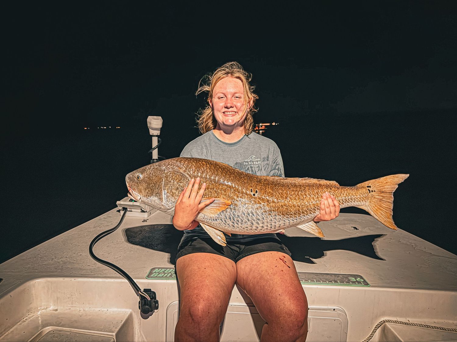 Smiling angler holds a large redfish on a boat at night during a successful fishing trip.