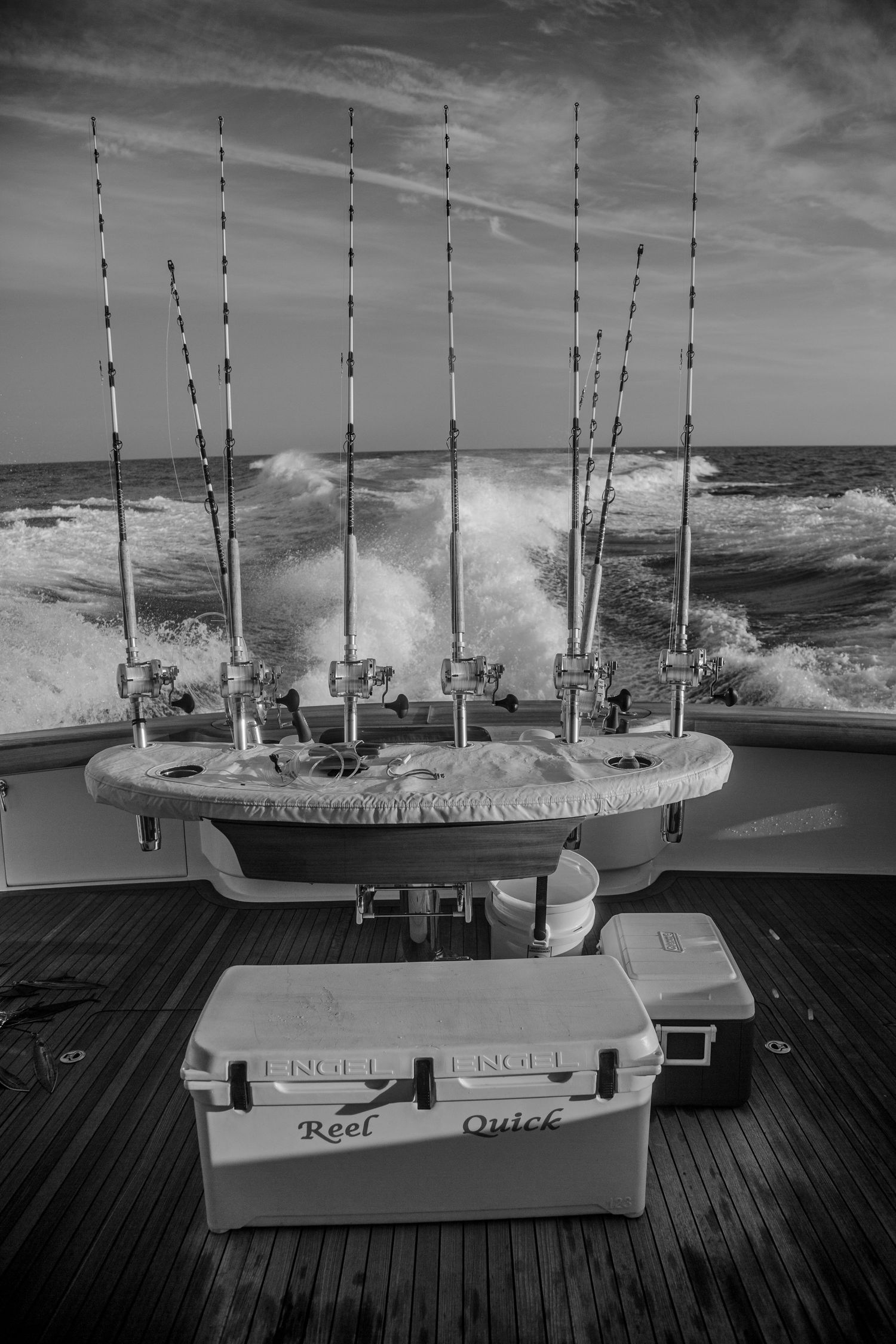 Black and white photo of deep sea fishing rods on a boat deck with YETI coolers labeled Reel Quick in rough ocean waters.
