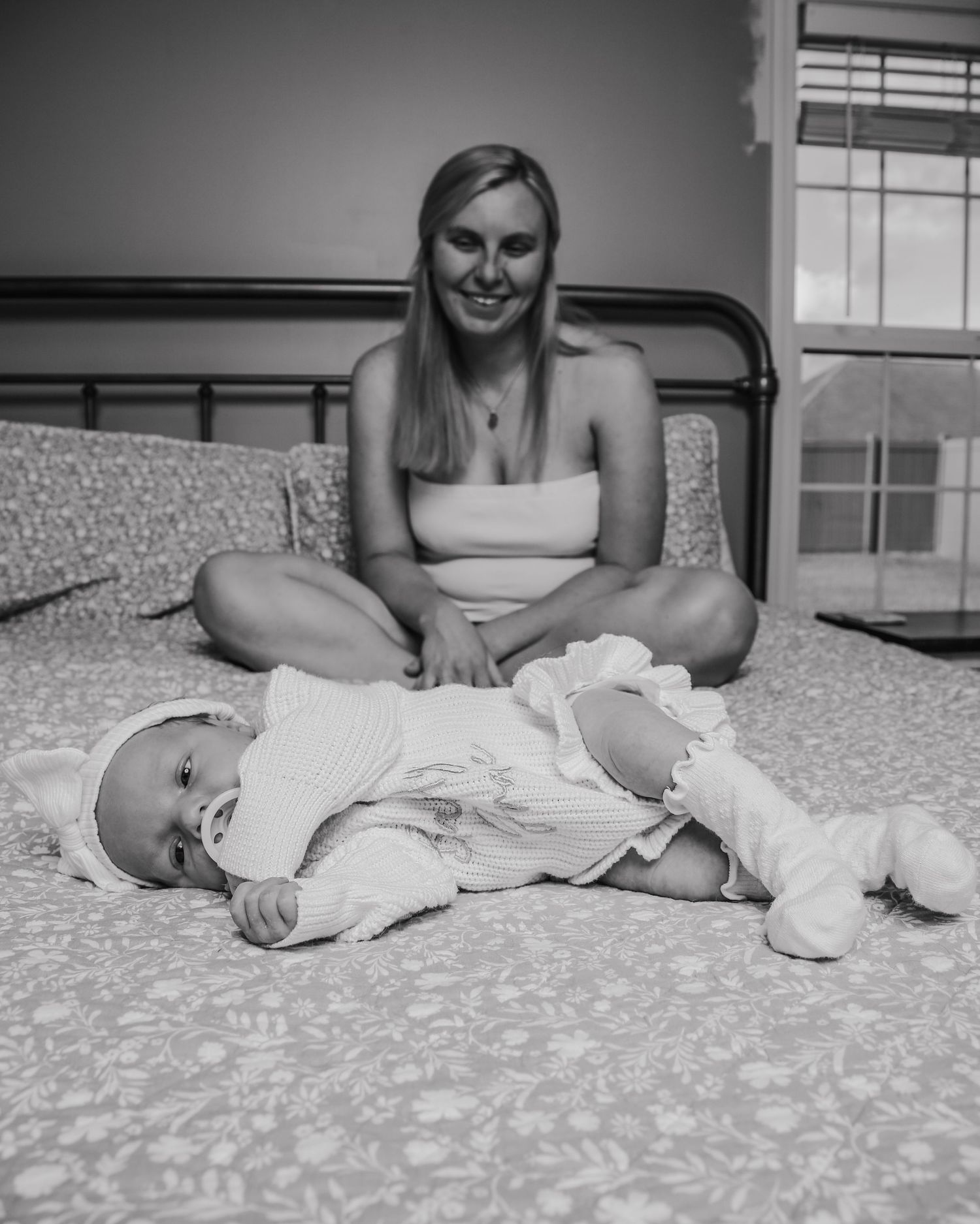 Black and white portrait of someone sitting cross-legged on a bed next to a baby laying on floral bedding.