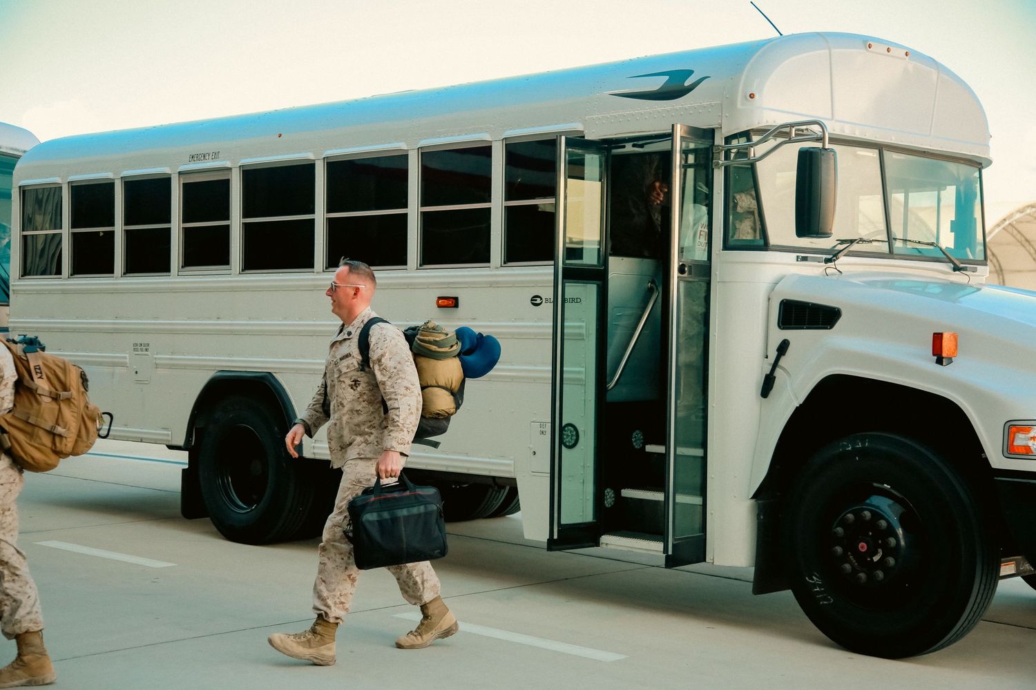 Military service member boarding a white school bus with luggage at a base or deployment location.