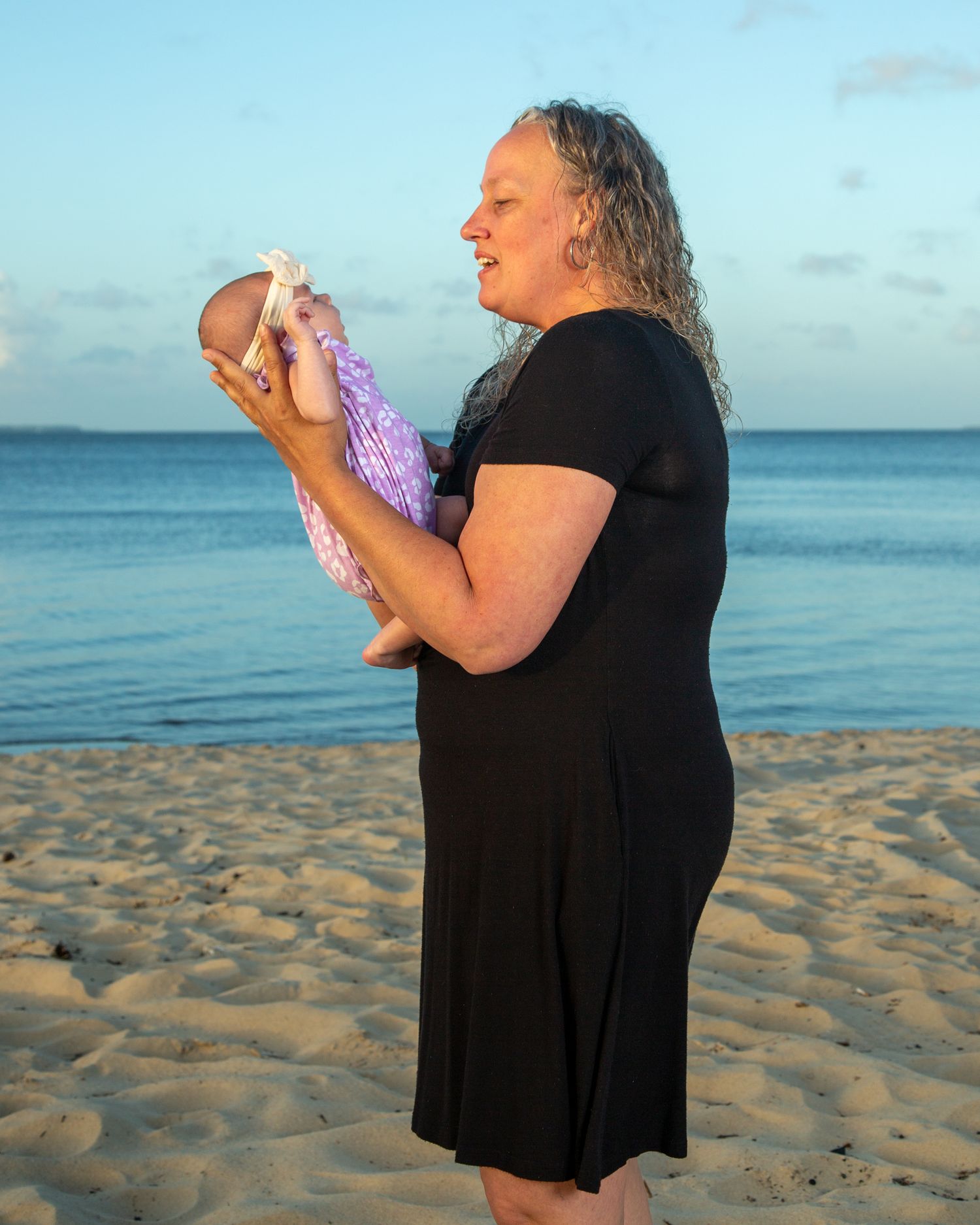 Woman in black dress holds newborn baby on sandy beach at sunset with calm blue ocean in background.