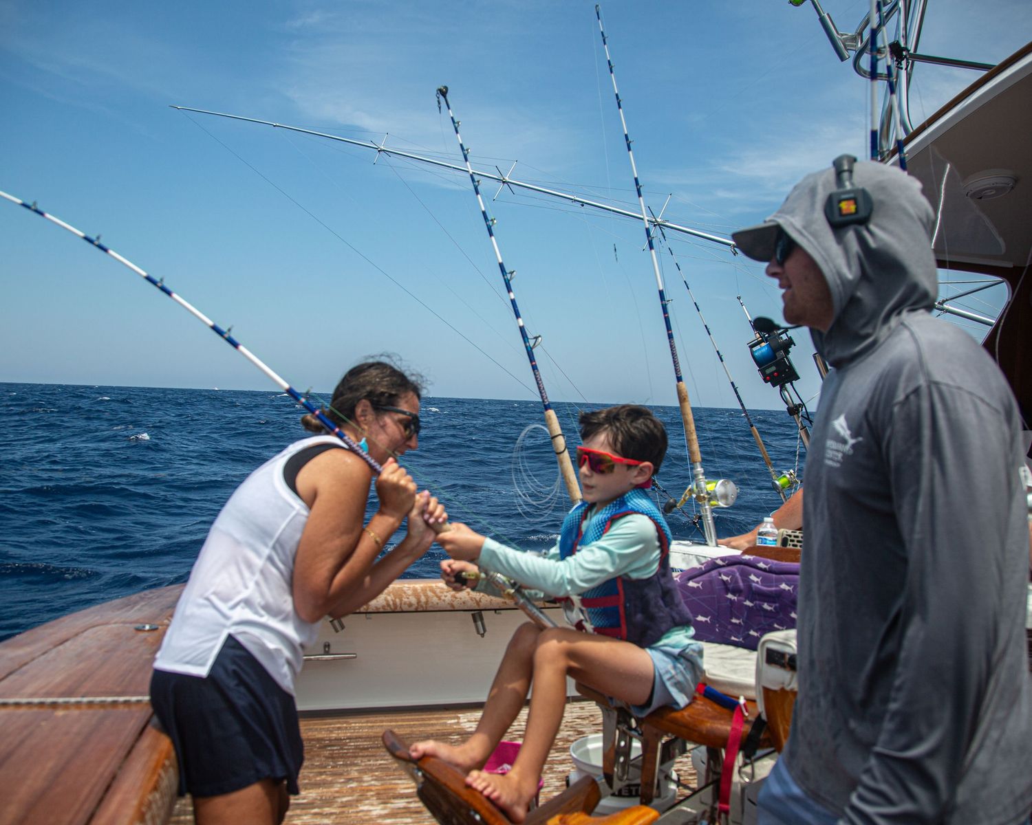 Family deep sea fishing on a boat, woman and child reeling in a catch while a man in a hoodie watches on a sunny day.