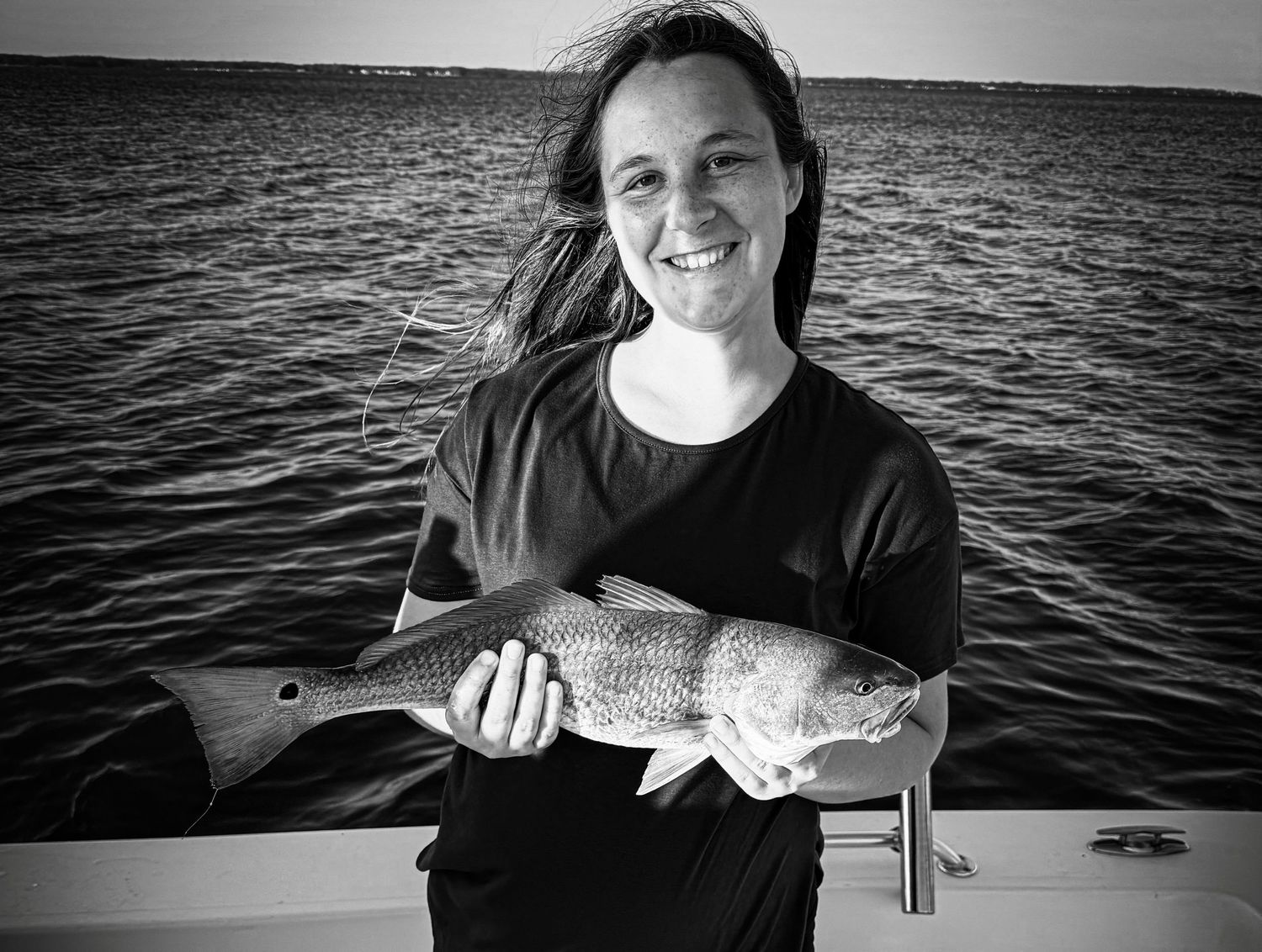 Woman smiling on a boat holding a large redfish catch over open water in black and white photo.