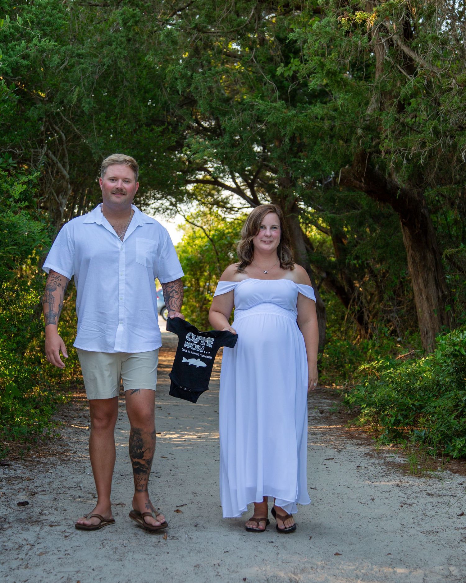 Pregnant couple in white outfits holding a baby onesie on a scenic tree-lined path during a maternity photo shoot.