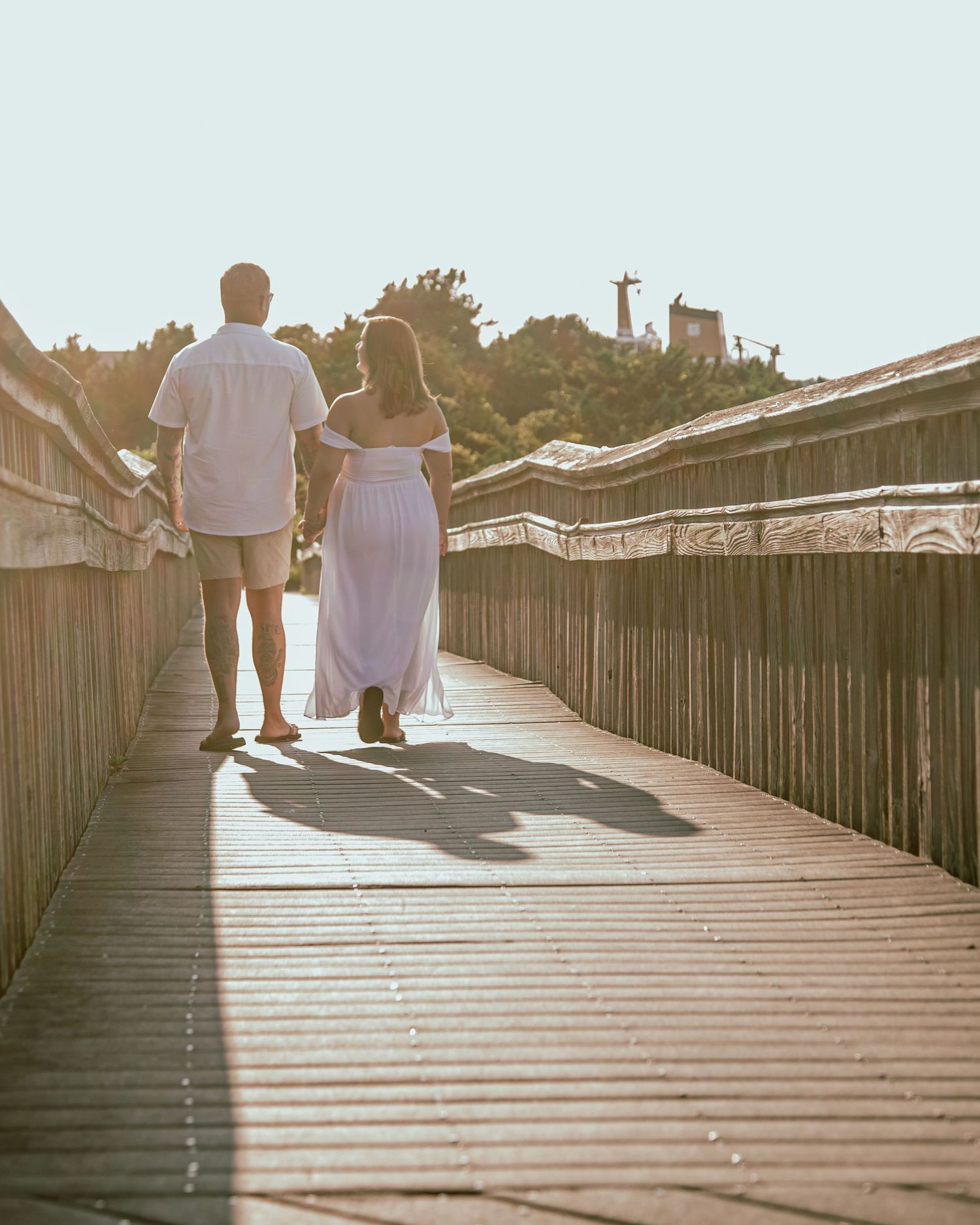 Couple walking hand-in-hand on a sunlit wooden boardwalk, man in white shirt and woman in pink maxi dress.