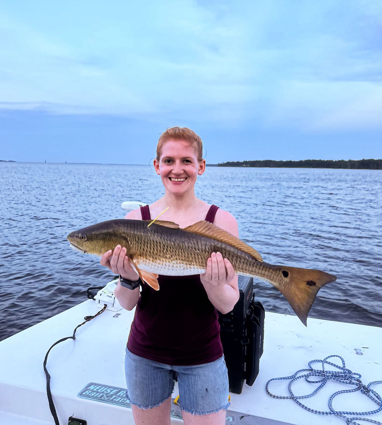 Angler displaying a large redfish catch on a boat during sunset over calm waters.