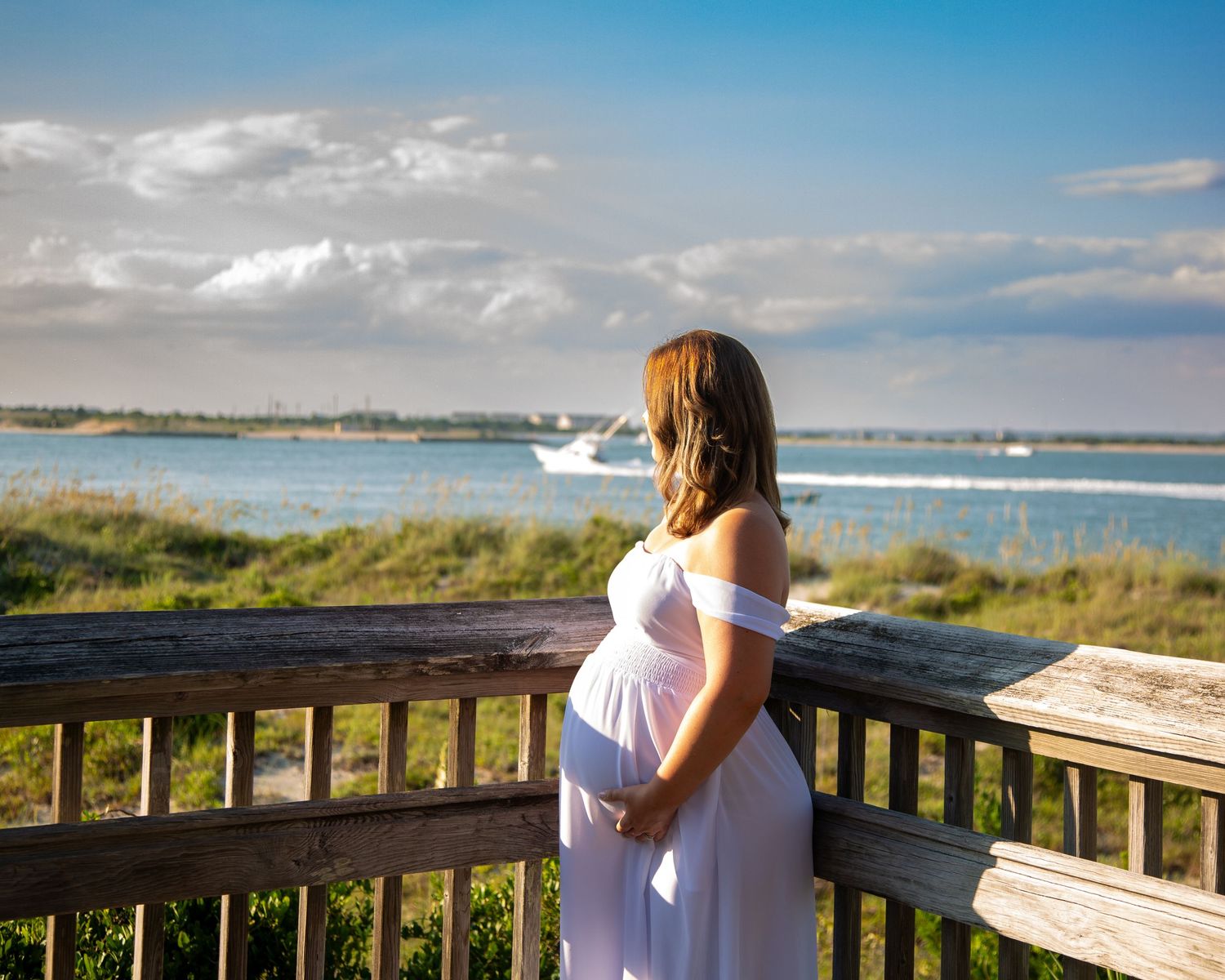 Pregnant woman in white dress stands on wooden deck overlooking calm coastal waters with a boat passing by.