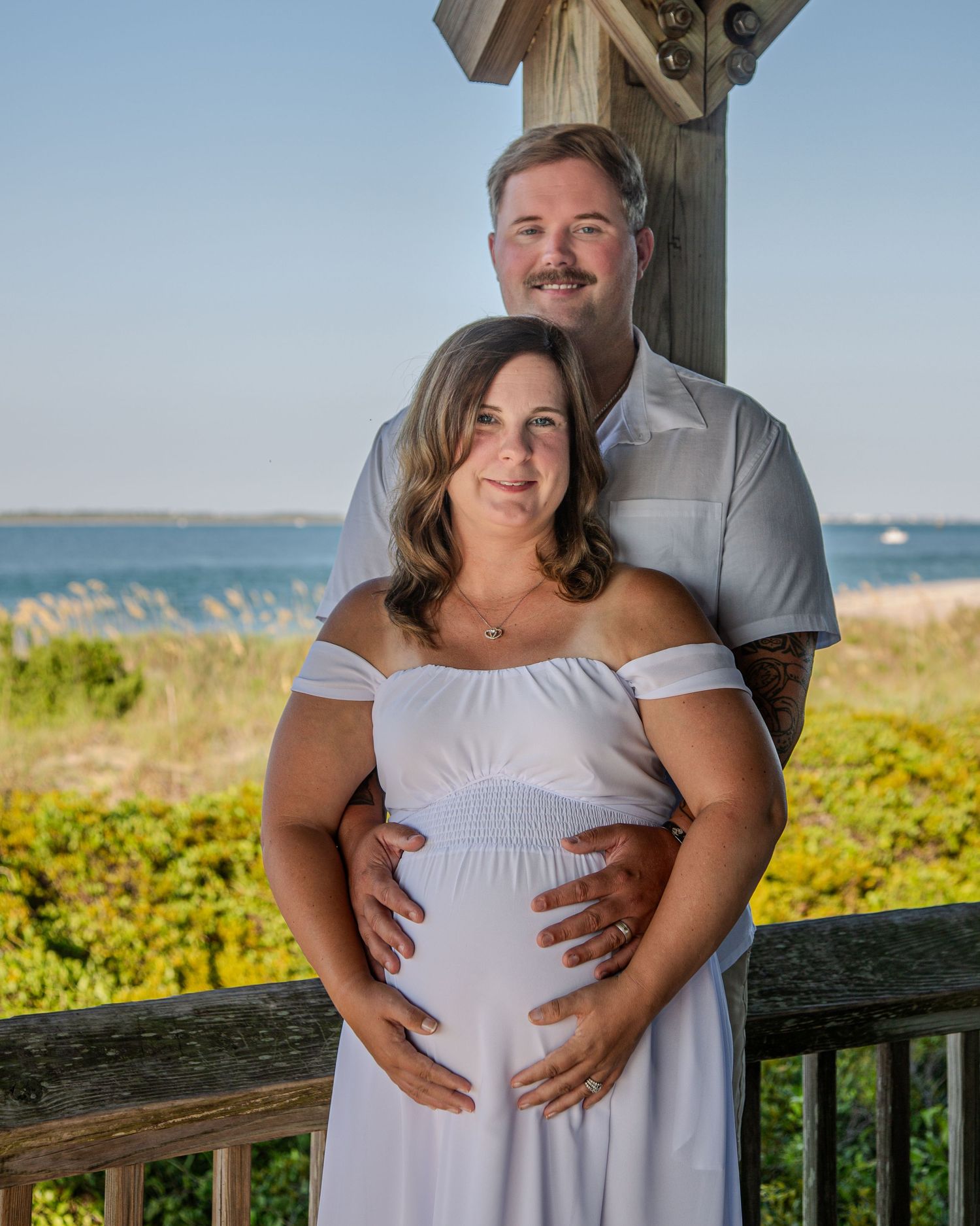 Couple posing for maternity photo shoot on beach deck, woman in white off-shoulder dress, sunny day.