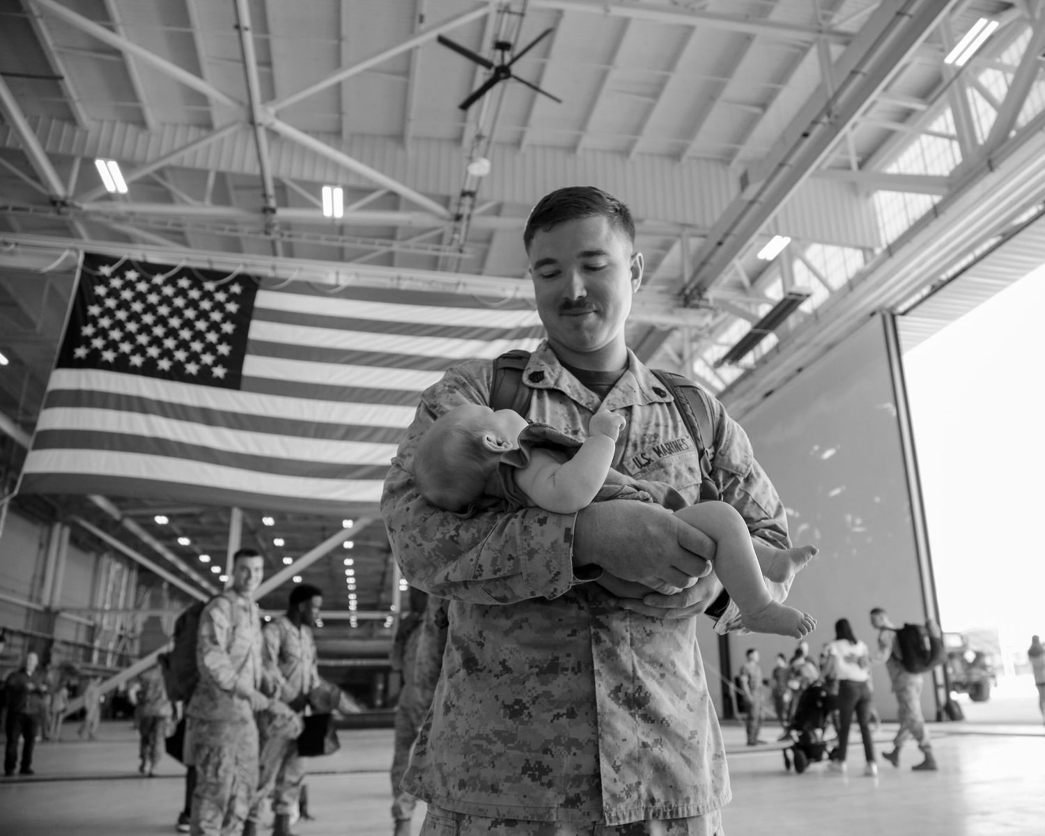 Military soldier in camouflage uniform holds infant under American flag in hangar homecoming ceremony.