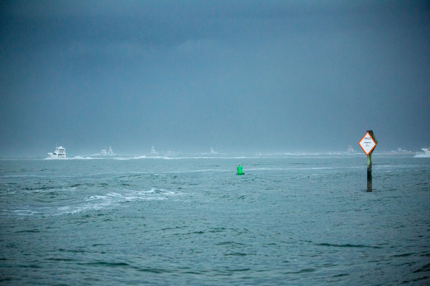 Distant boats navigate past channel markers in stormy seas under gray skies.