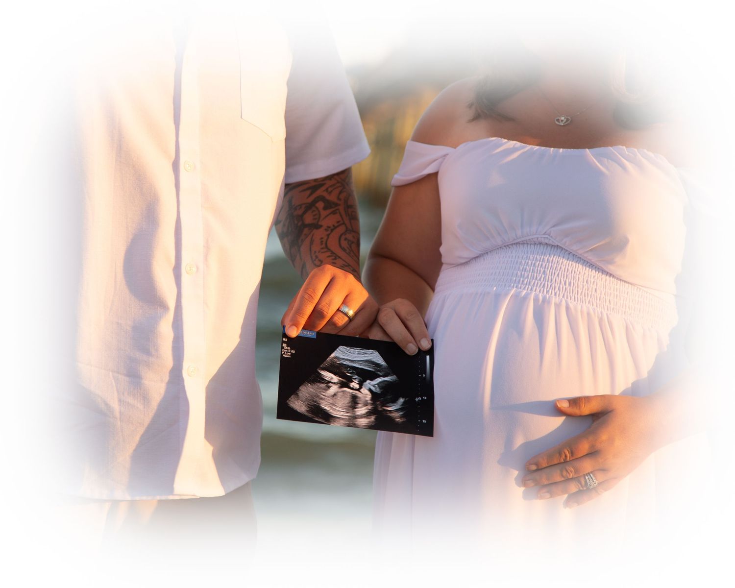 Pregnant couple in white outfits holding an ultrasound photo outdoors, celebrating their upcoming baby.