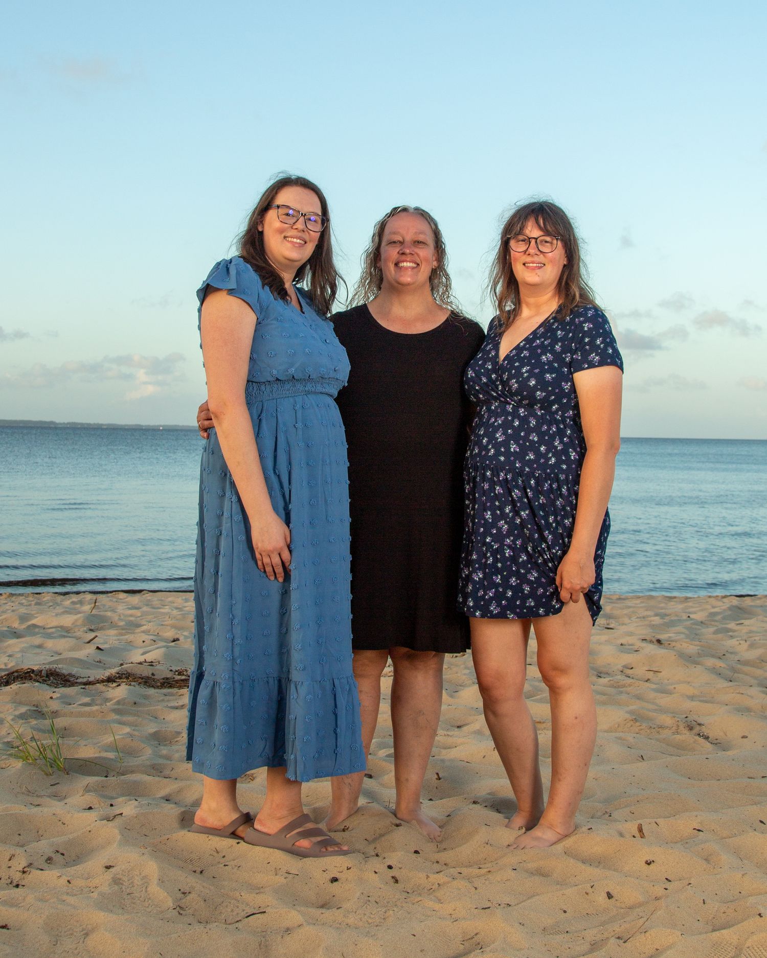 Three women smiling together on a sandy beach at sunset, wearing casual dresses, with ocean waves in the background.