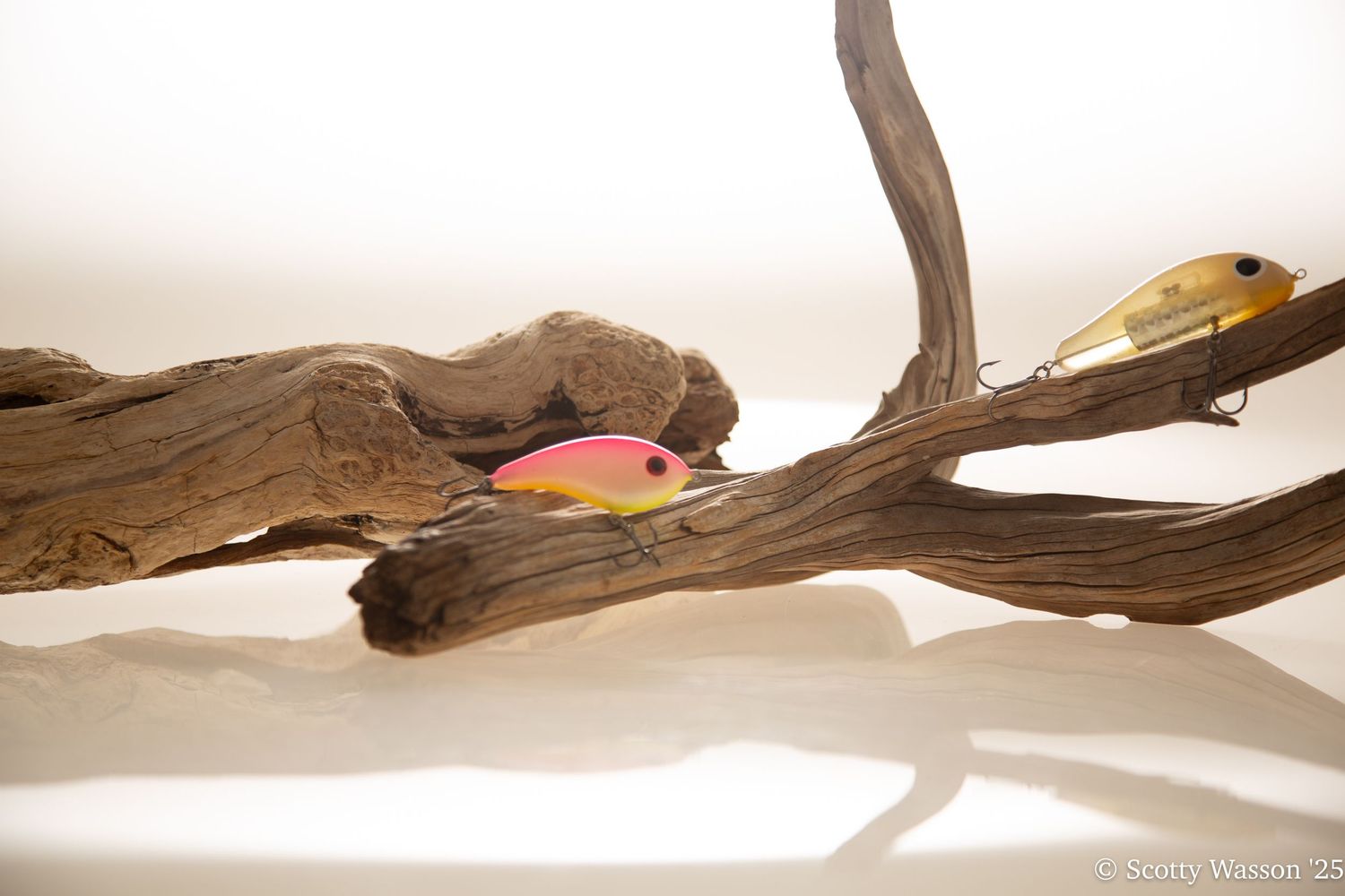 Two colorful fishing lures rest on driftwood against a white background, reflecting on a glossy surface.