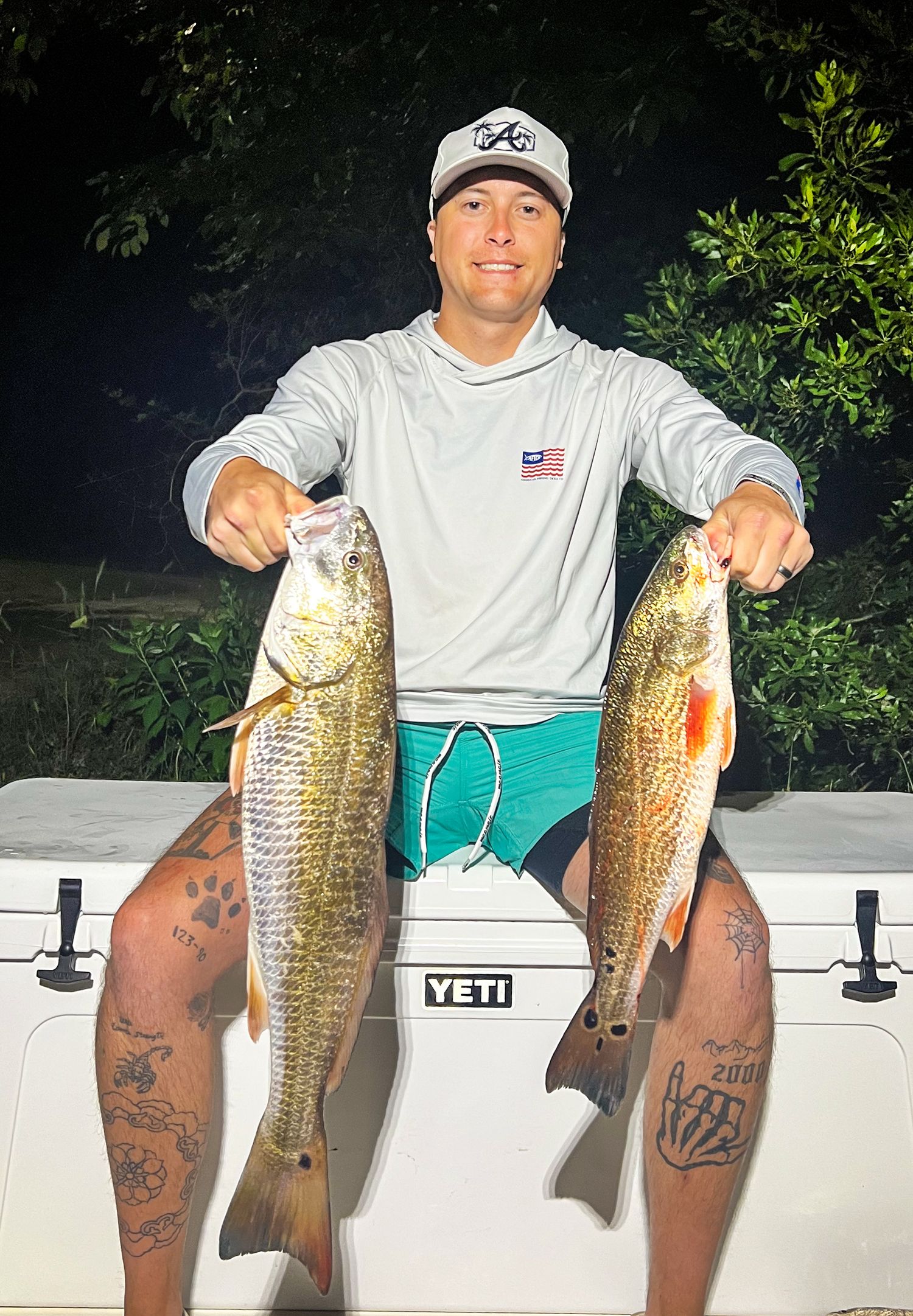 Man holding two large redfish while sitting on a Yeti cooler at night after a successful fishing trip.