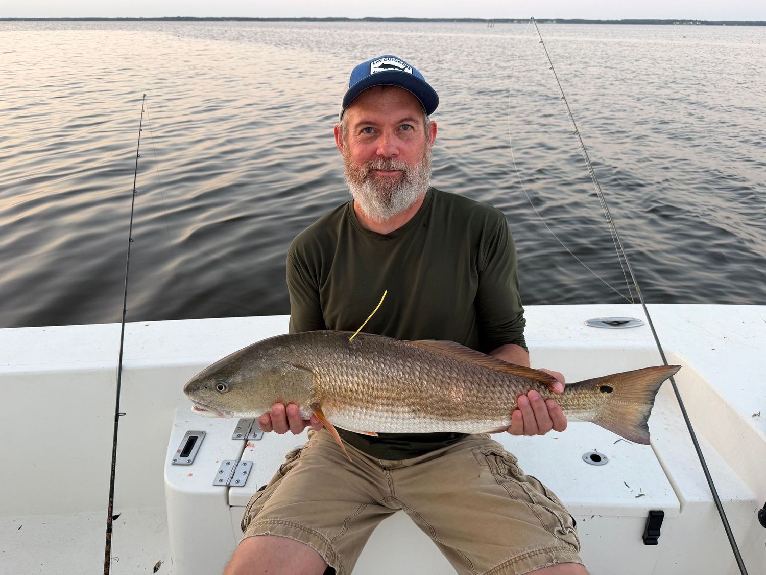 Man in blue cap holds a large redfish on a boat at dusk on calm open water.