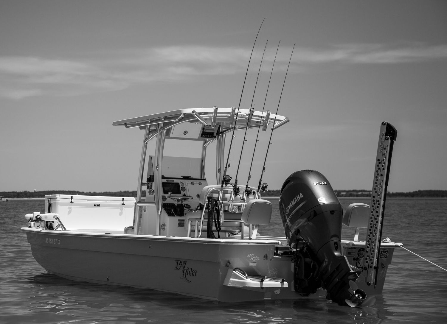 A white fishing boat with fishing rods and an outboard motor anchored on calm waters in black and white photography.