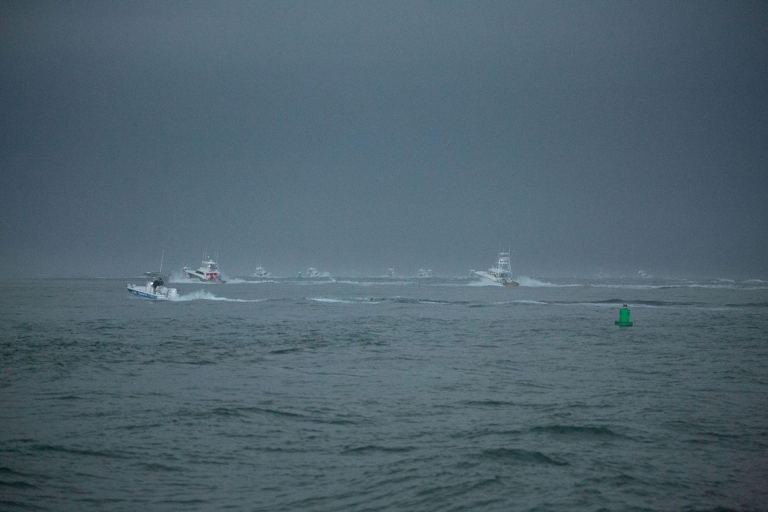 Multiple boats navigate through choppy waters with a green channel marker bobbing in the waves.
