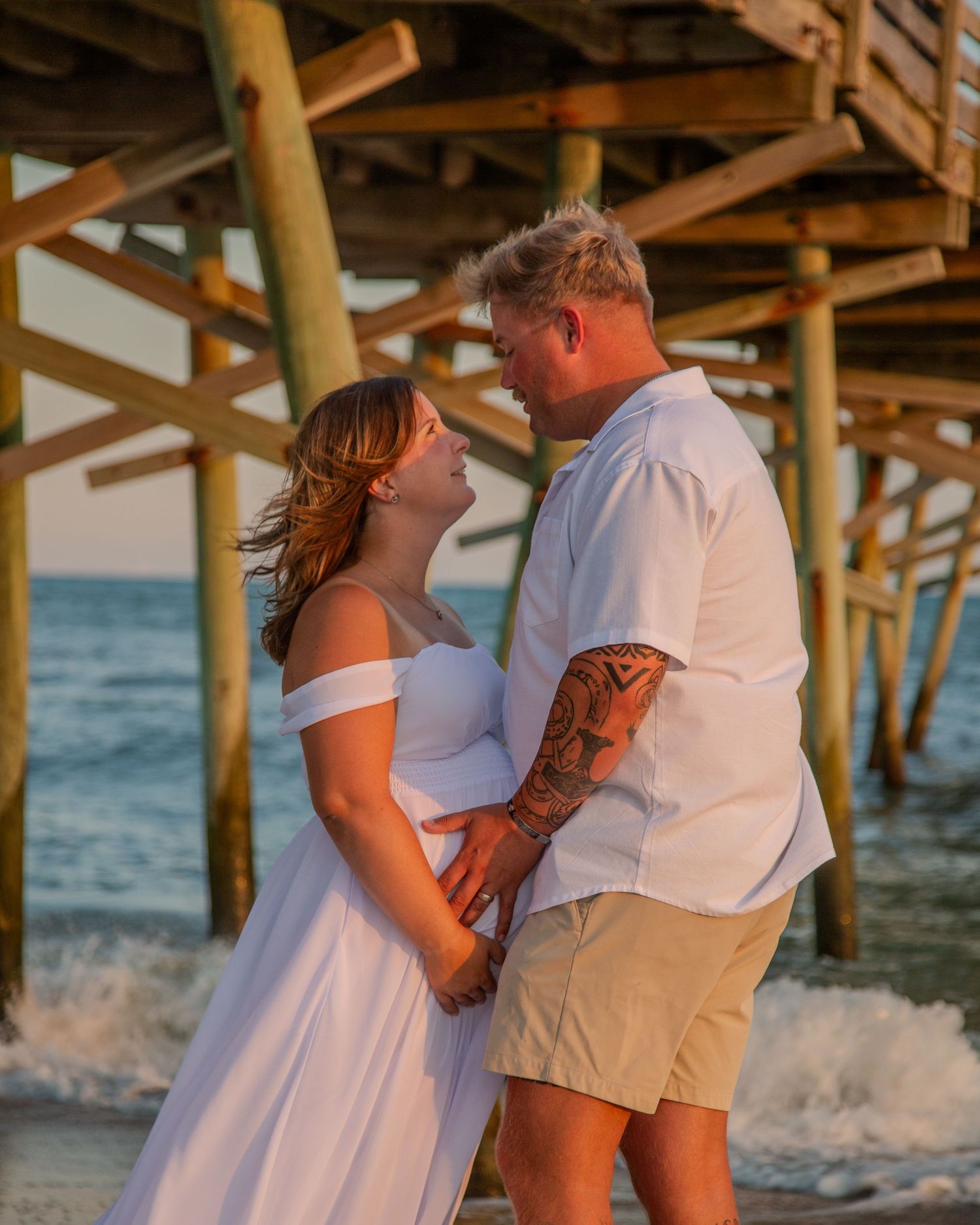 Couple in white outfits holding hands under a pier on the beach at sunset, ocean waves in the background.