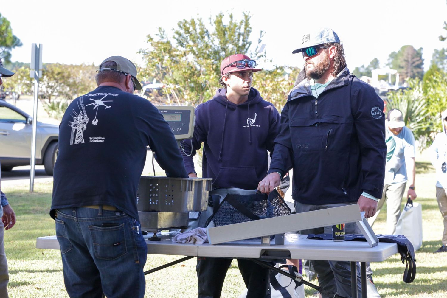 Three people examining fishing equipment on display table at outdoor event.