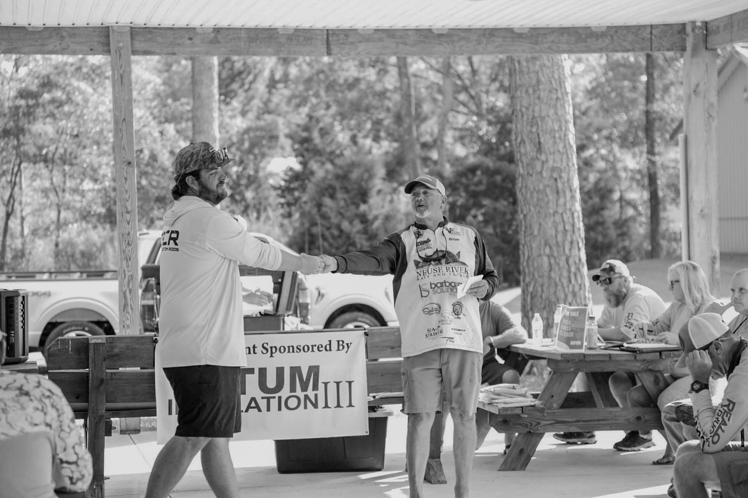 Black and white image of outdoor event with people gathered around picnic tables.