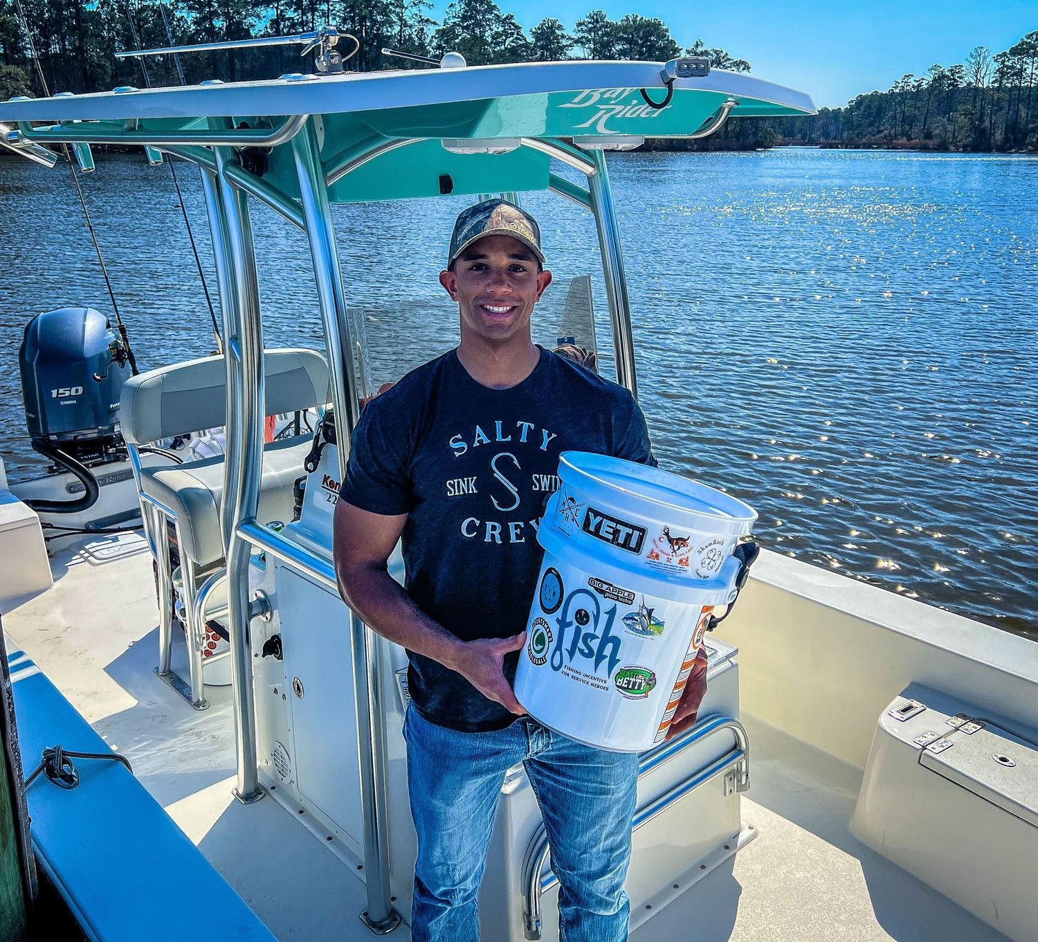 Man in Salty Crew shirt holding a fish bucket on a center console boat on calm water.