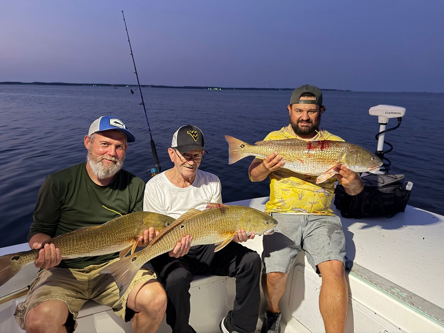 Three anglers proudly displaying large redfish caught on a fishing boat at dusk.