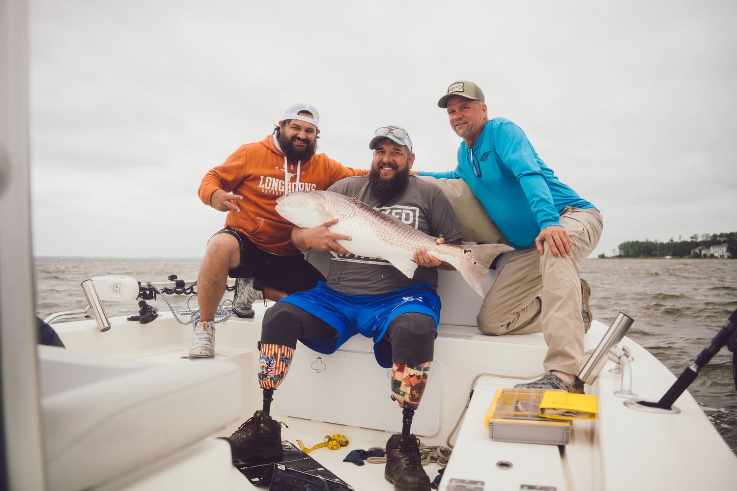 Three men on a boat proudly display a large redfish catch, one man with prosthetic legs, on a cloudy fishing trip.