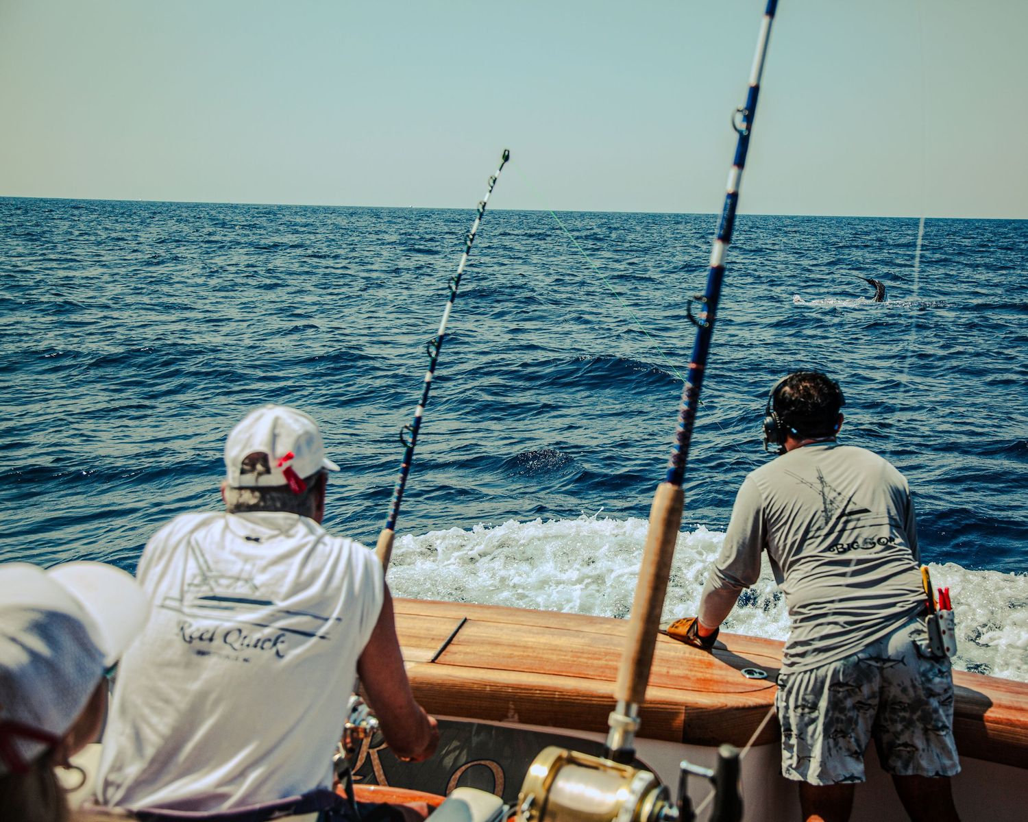 Two anglers fishing from the stern of a sport fishing boat in open blue ocean waters.