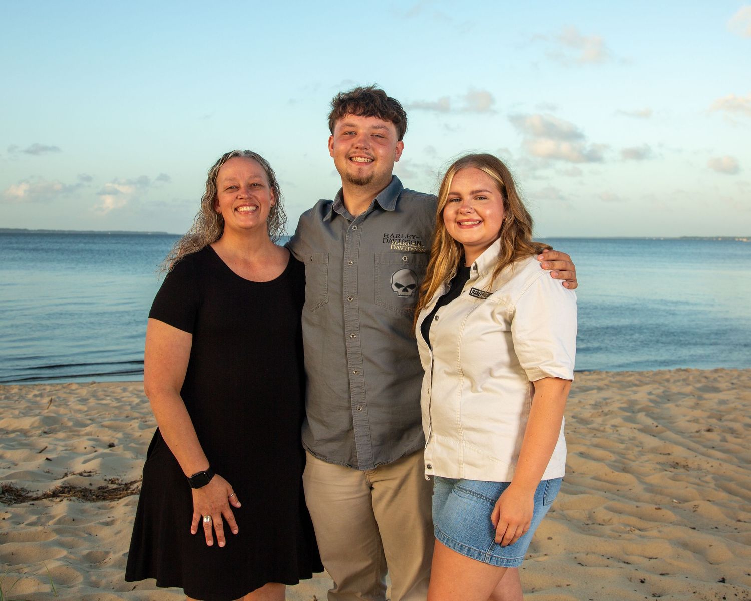 Three smiling people pose together on a sandy beach at sunset with calm ocean waters in the background.