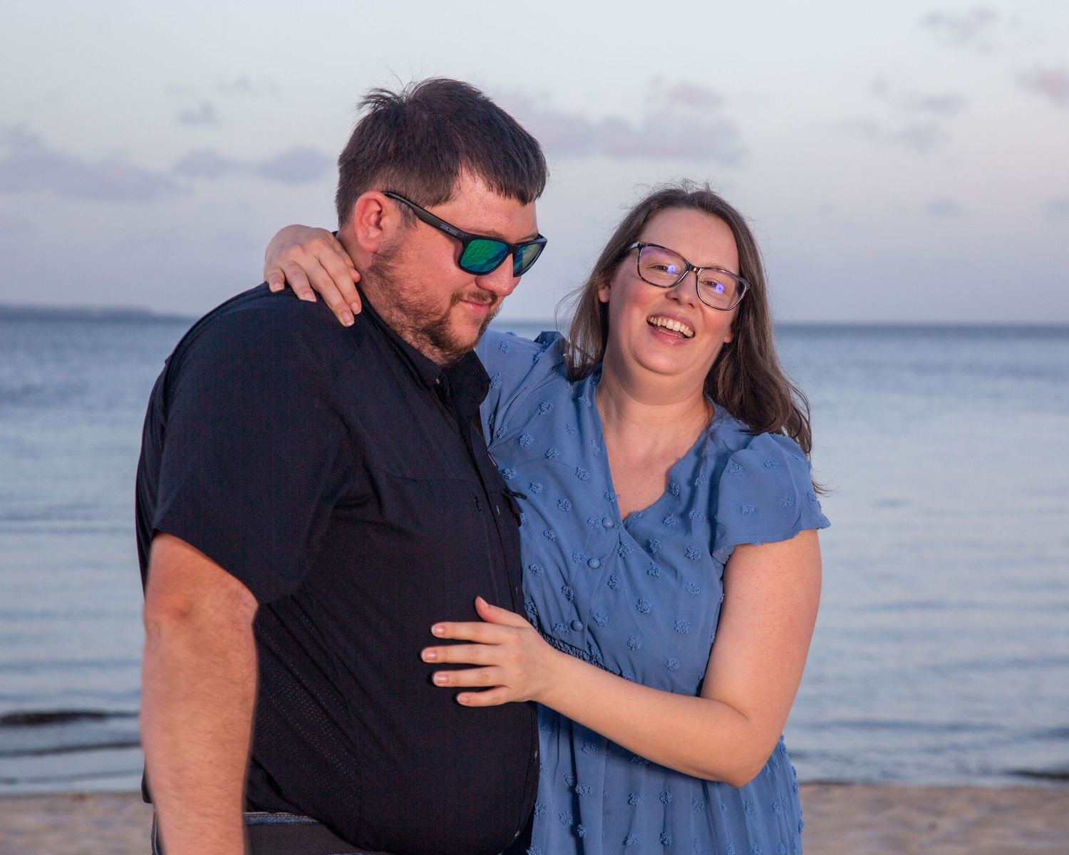 Couple smiling and embracing on a beach at sunset, both wearing glasses, woman in blue dress, man in black shirt.