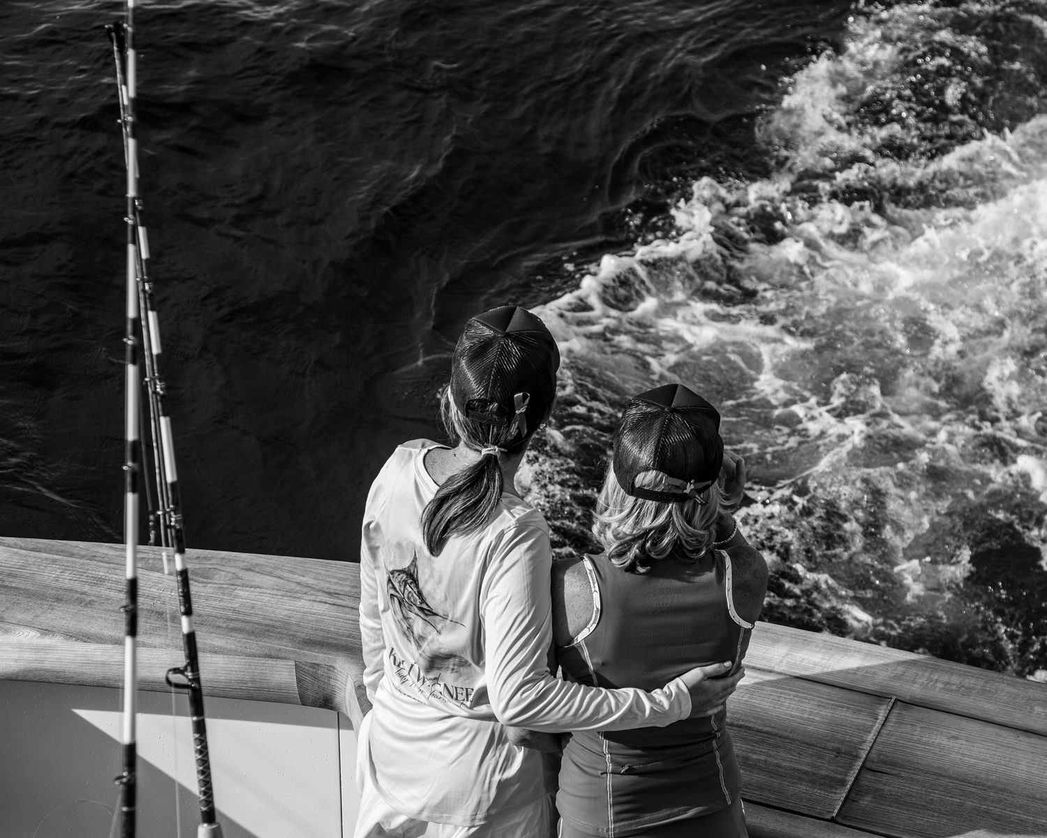 Two women lean over the railing of a boat watching ocean waves in a black and white photo.
