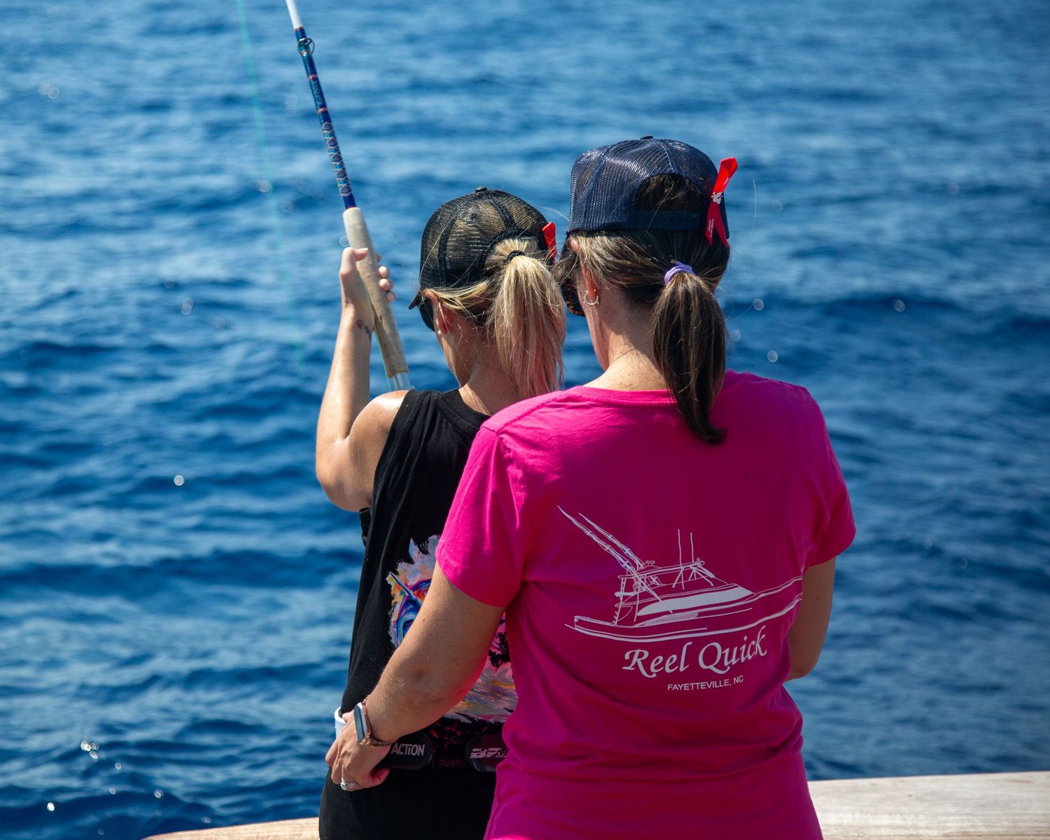 Two women fishing off a pier, one wearing a pink Reel Quick shirt, with blue ocean water in the background.