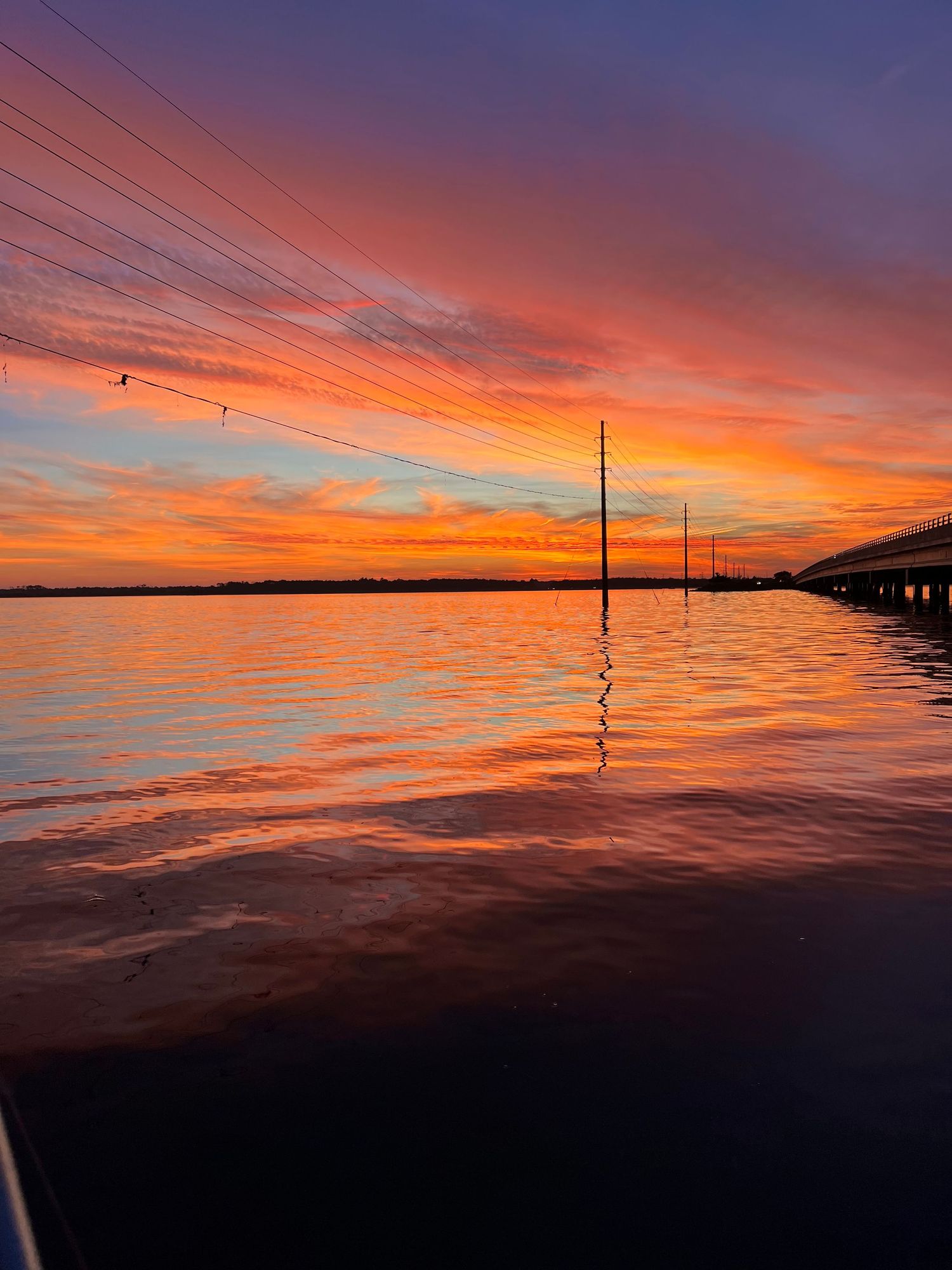 Vibrant orange and purple sunset sky reflected in water with power lines silhouetted against the horizon.