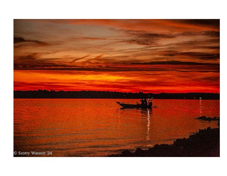 Dramatic red sunset over water with silhouetted shoreline and deep orange reflections.