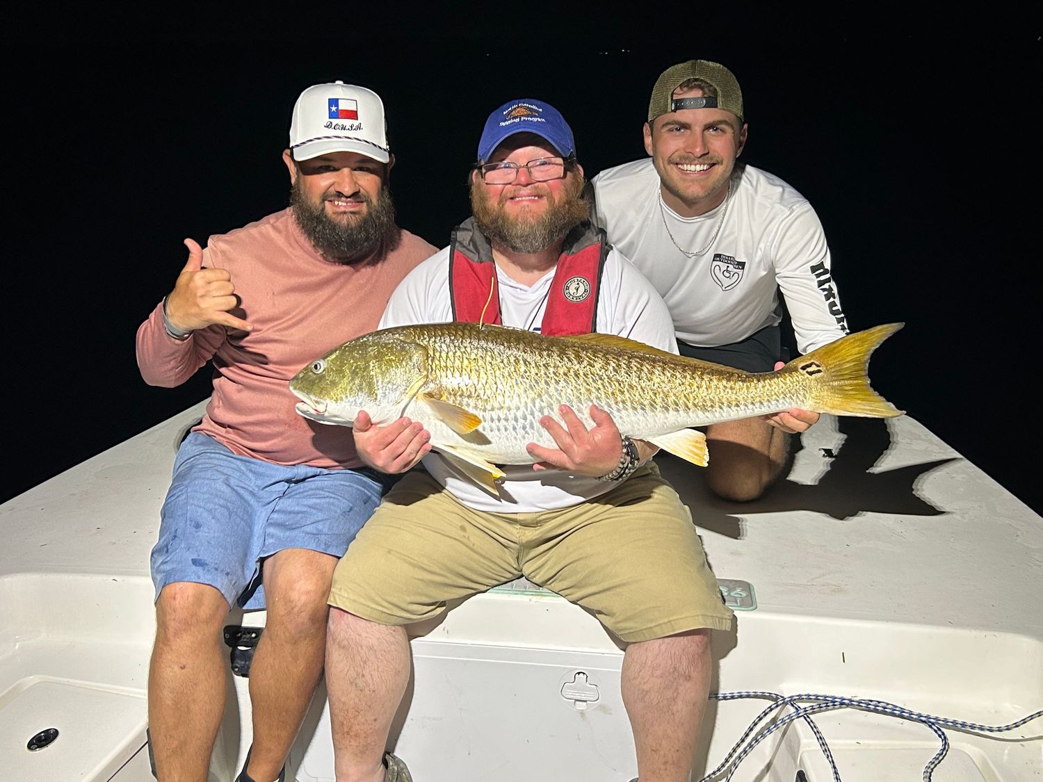 Three anglers displaying their impressive redfish catch on a boat at night.
