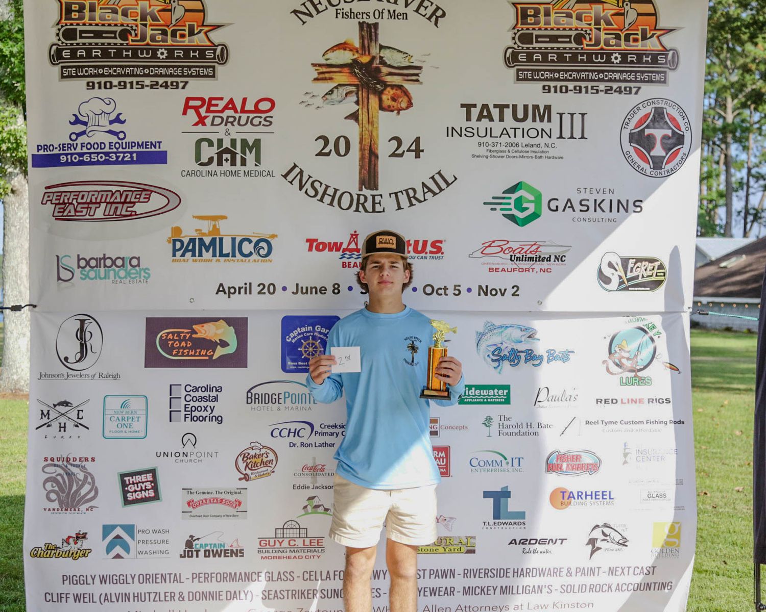 Person standing in front of sponsor banner wall at an outdoor fishing competition event.