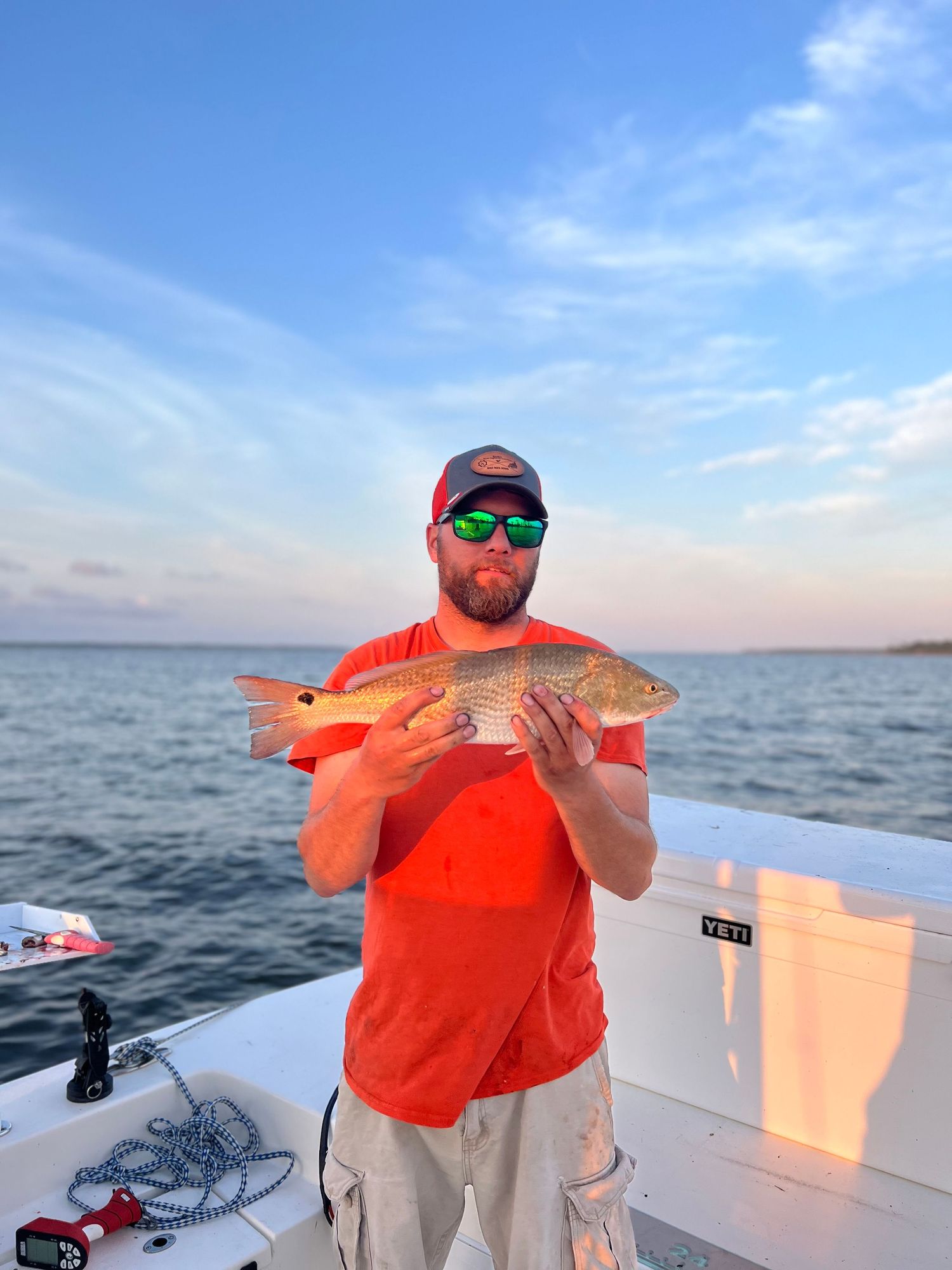 Fisherman in orange shirt holds up red snapper catch on boat during sunset fishing trip.