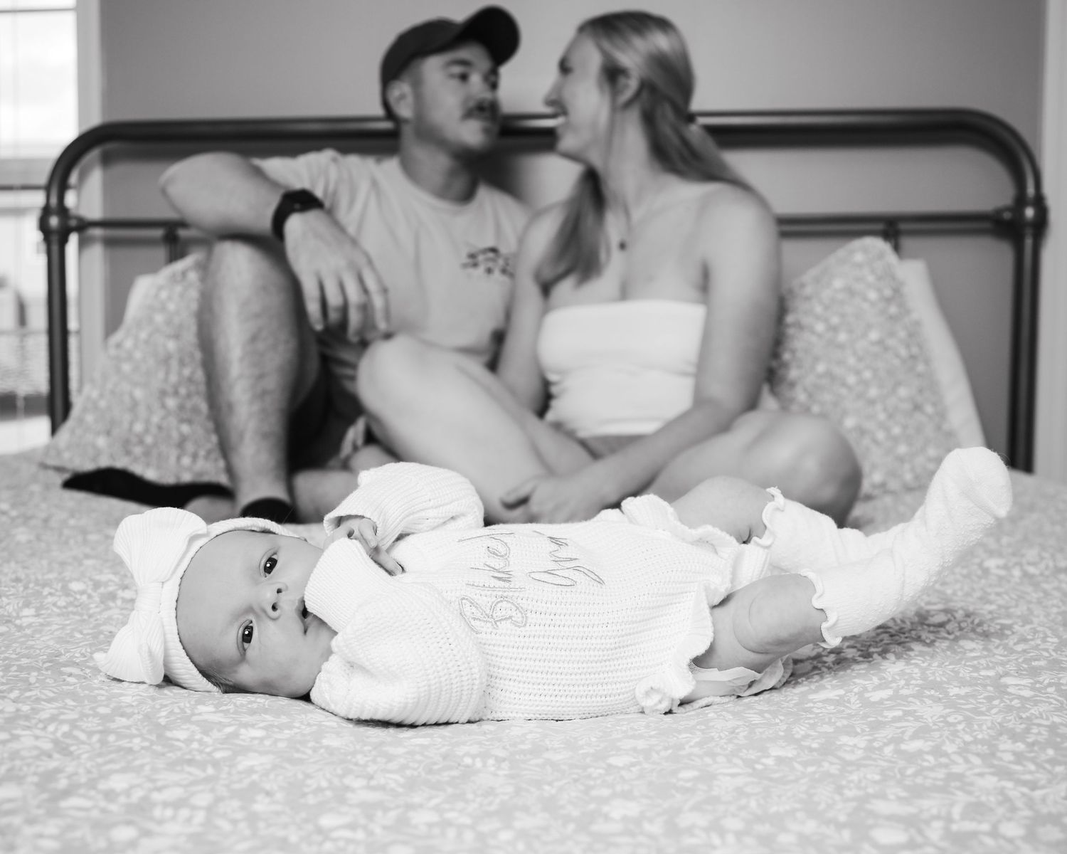Black and white photo of a baby lying on bed in white clothes while a couple sits together in the background.