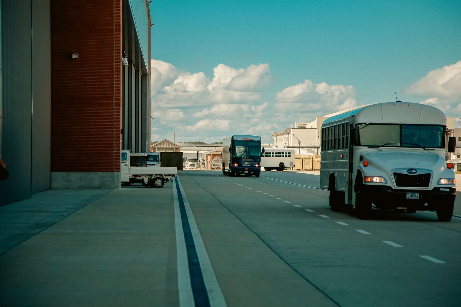 Row of industrial loading docks under bright blue sky with white puffy clouds and parked trucks in distance.