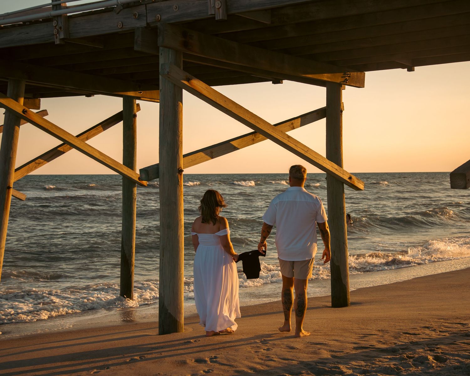 Couple holding hands at sunset under a wooden beach pier, waves crashing on sandy shore in warm golden light.