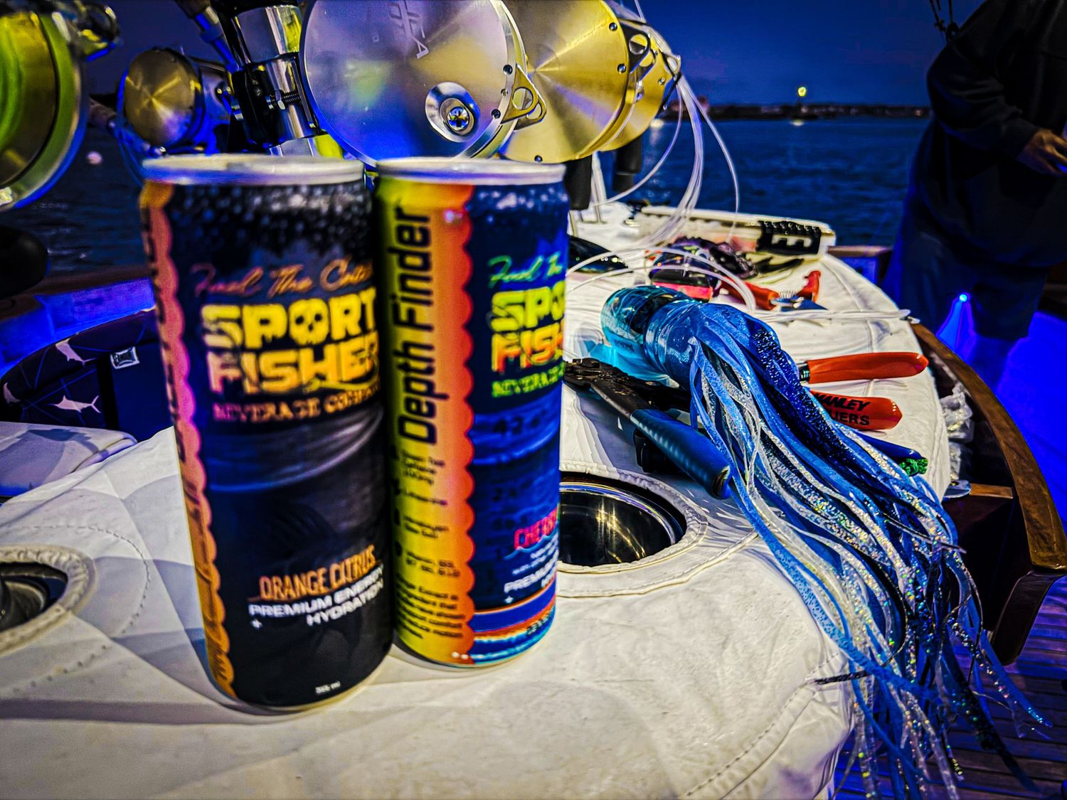 Sport Fisher energy drink cans on a fishing boat at night with blue fishing lures and tackle visible in the background.